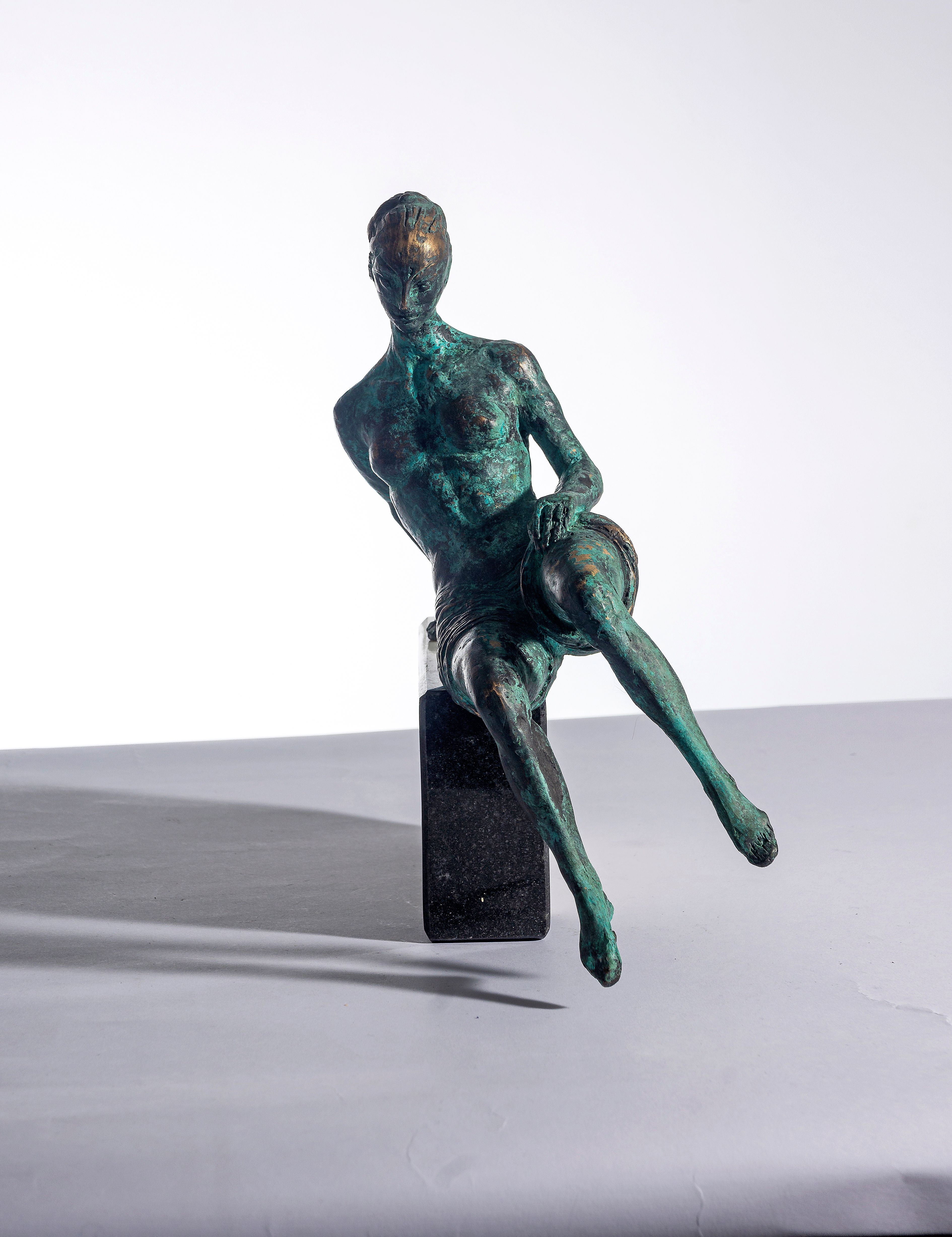 Bronze sculpture “Anna”: a small seated nude woman with one leg stretched forward, green-brown patina, sitting on a black stone block.