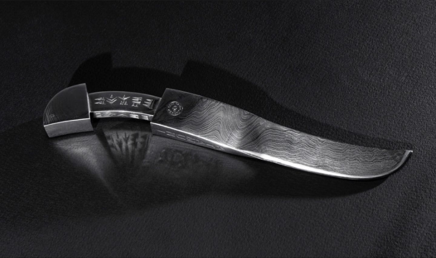Damascus steel composition “ATAMUNI”: a knife with a rippled blade and a handle bearing a row of small wedge-shaped signs, on a dark cloth.