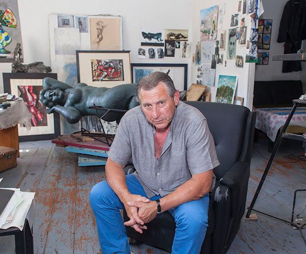 Gena Lakoba sitting in an armchair in his studio, wearing a grey shirt and blue trousers, with a bronze reclining figure and pinned drawings and paintings on the wall behind him.