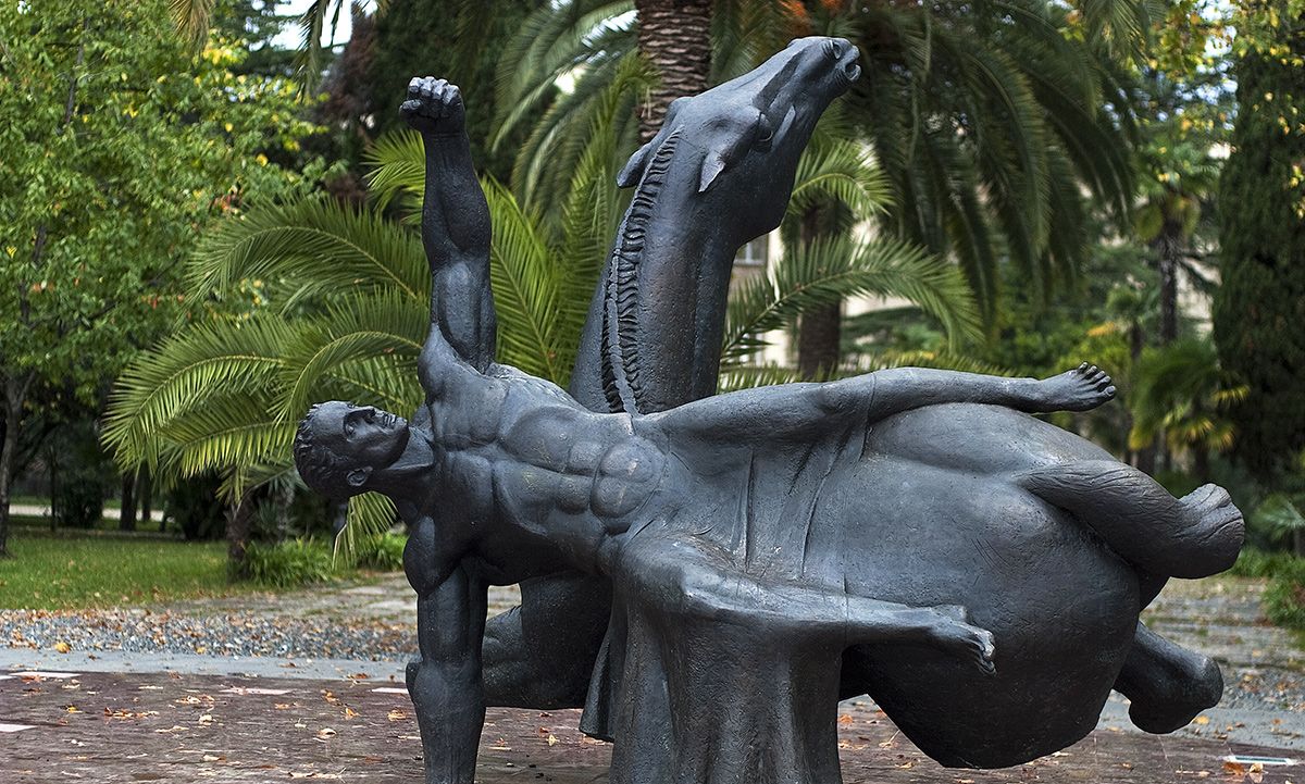 Dark bronze sculpture of a muscular man arching backwards beside a rearing horse, standing in a park among palm trees.