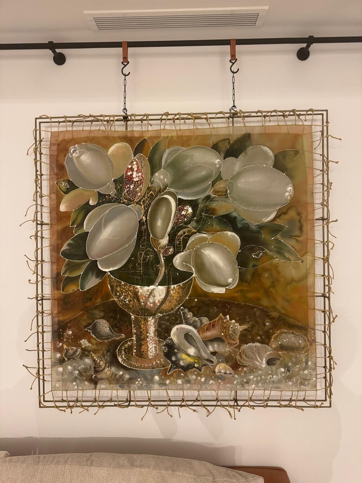 Square silk painting on a golden-brown background, laced with cords into a double metal frame: large silvery-white magnolia blossoms and dark leaves rise from a gold-speckled stemmed cup, with seashells scattered around its base.