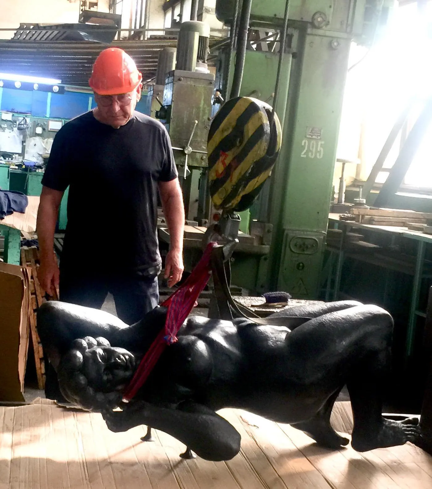 Sculptor in an orange hard hat looking at the black figure hanging from a hoist in a workshop