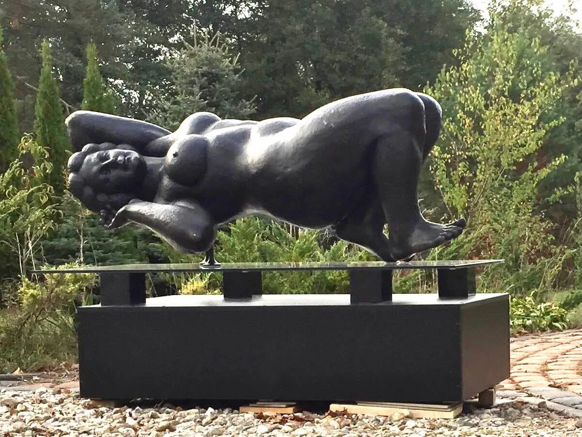 Black bronze Margo reclining on a glass sheet above a black plinth in a garden