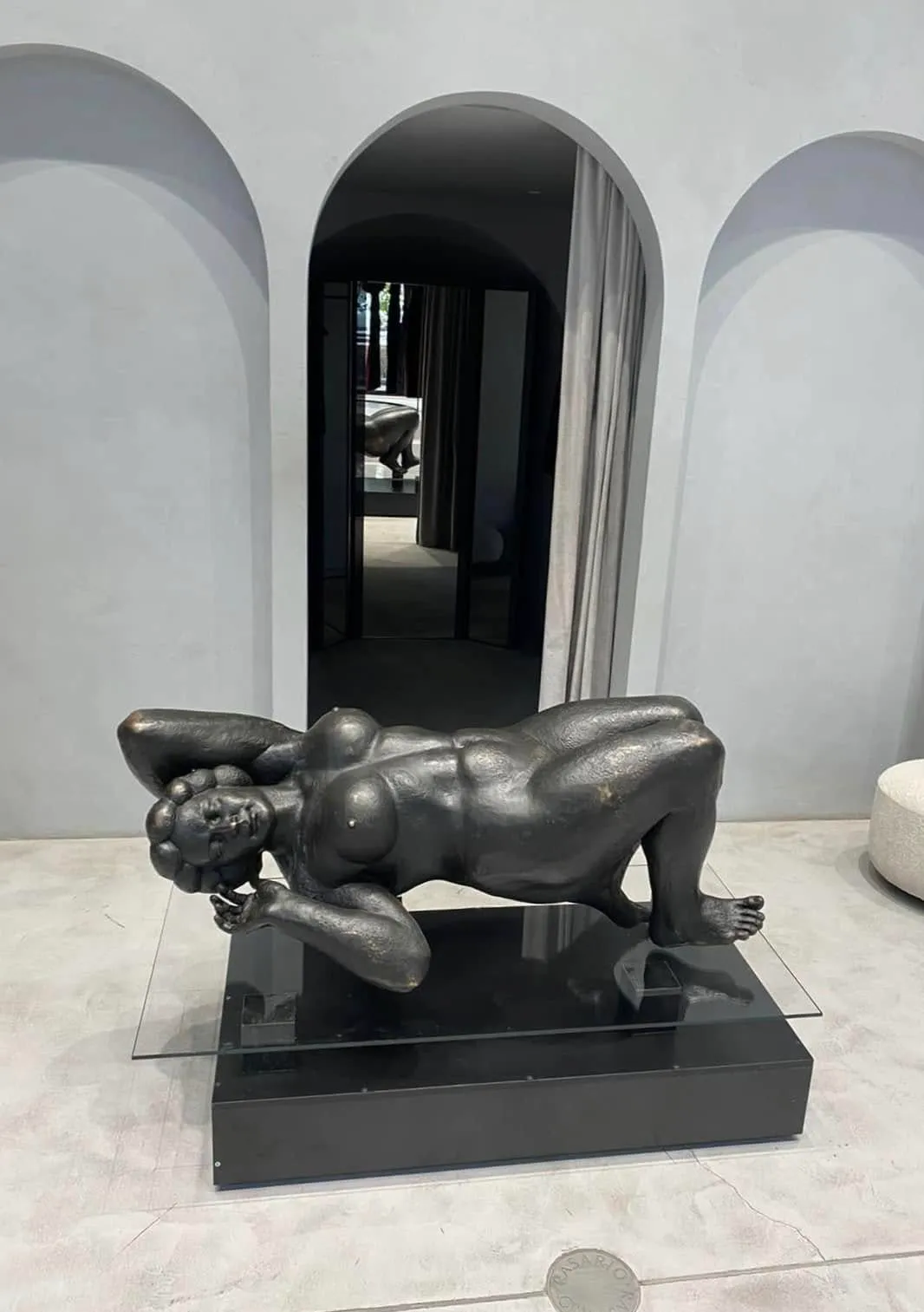 Margo reclining on a glass sheet over a black plinth in a gallery with arched grey walls