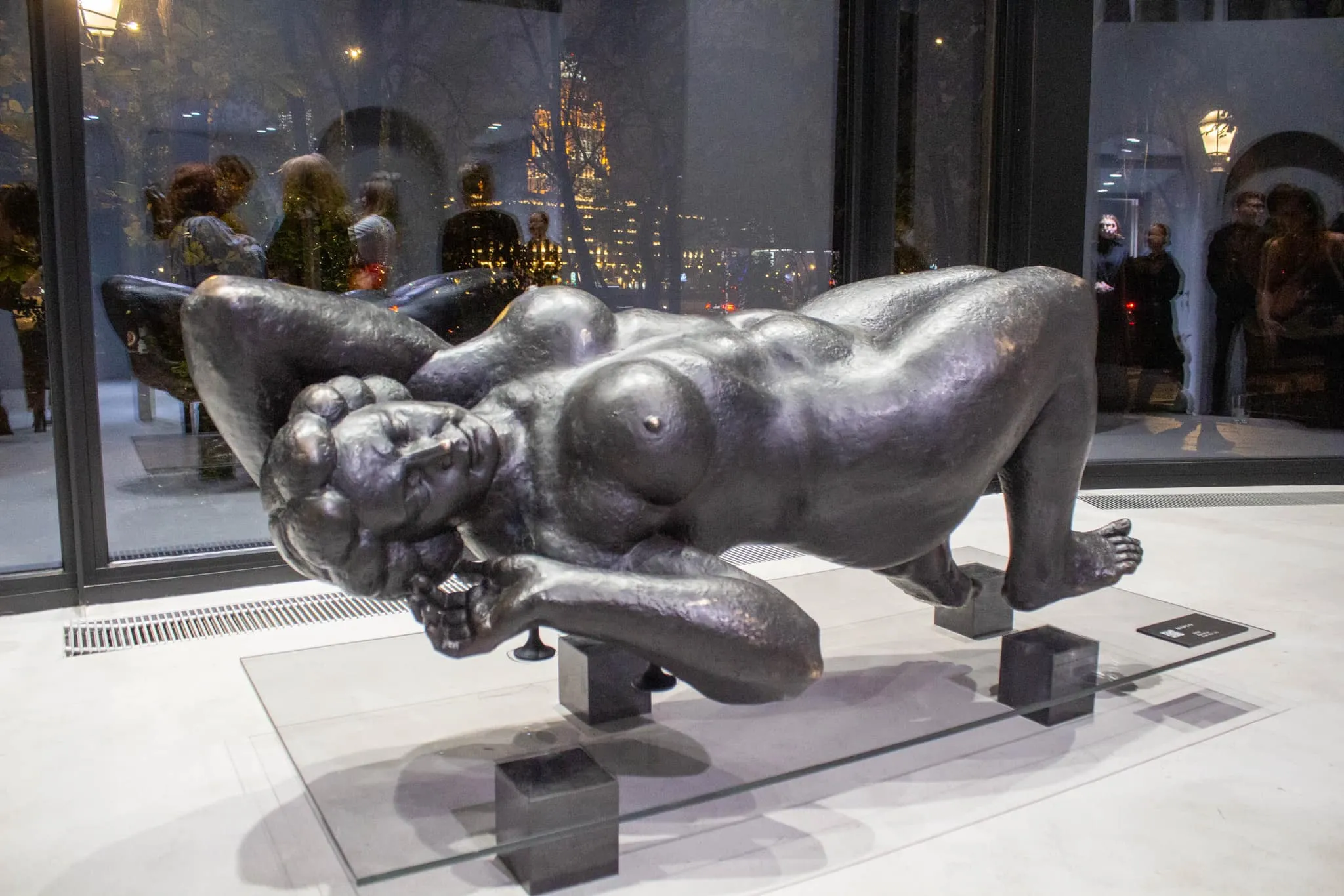 Margo displayed on a glass sheet and black cubes in front of a night-time window, with visitors behind