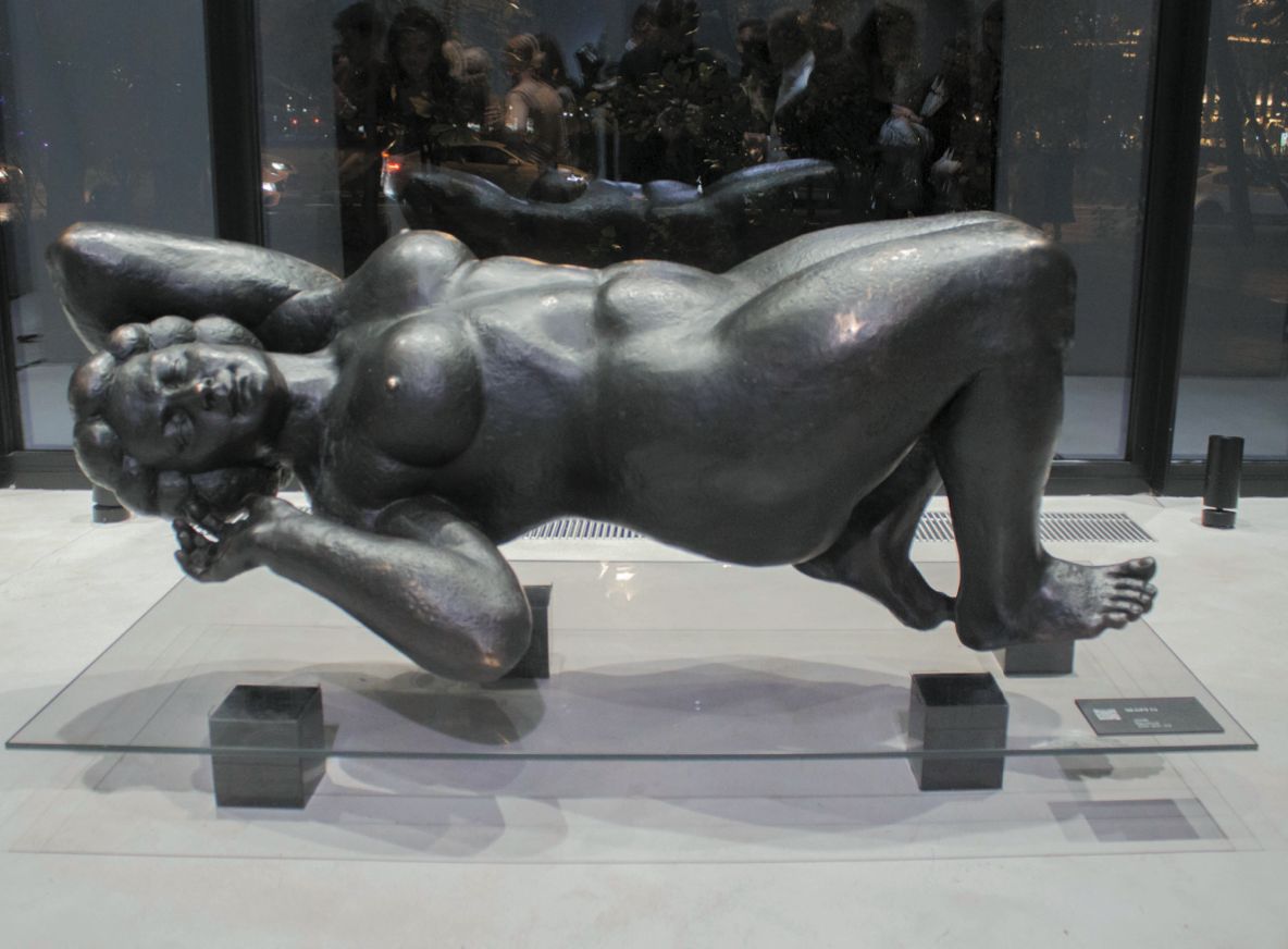 Bronze sculpture “Margo”: a large reclining nude woman, head propped on one hand, lying on a glass table inside a building with windows behind.