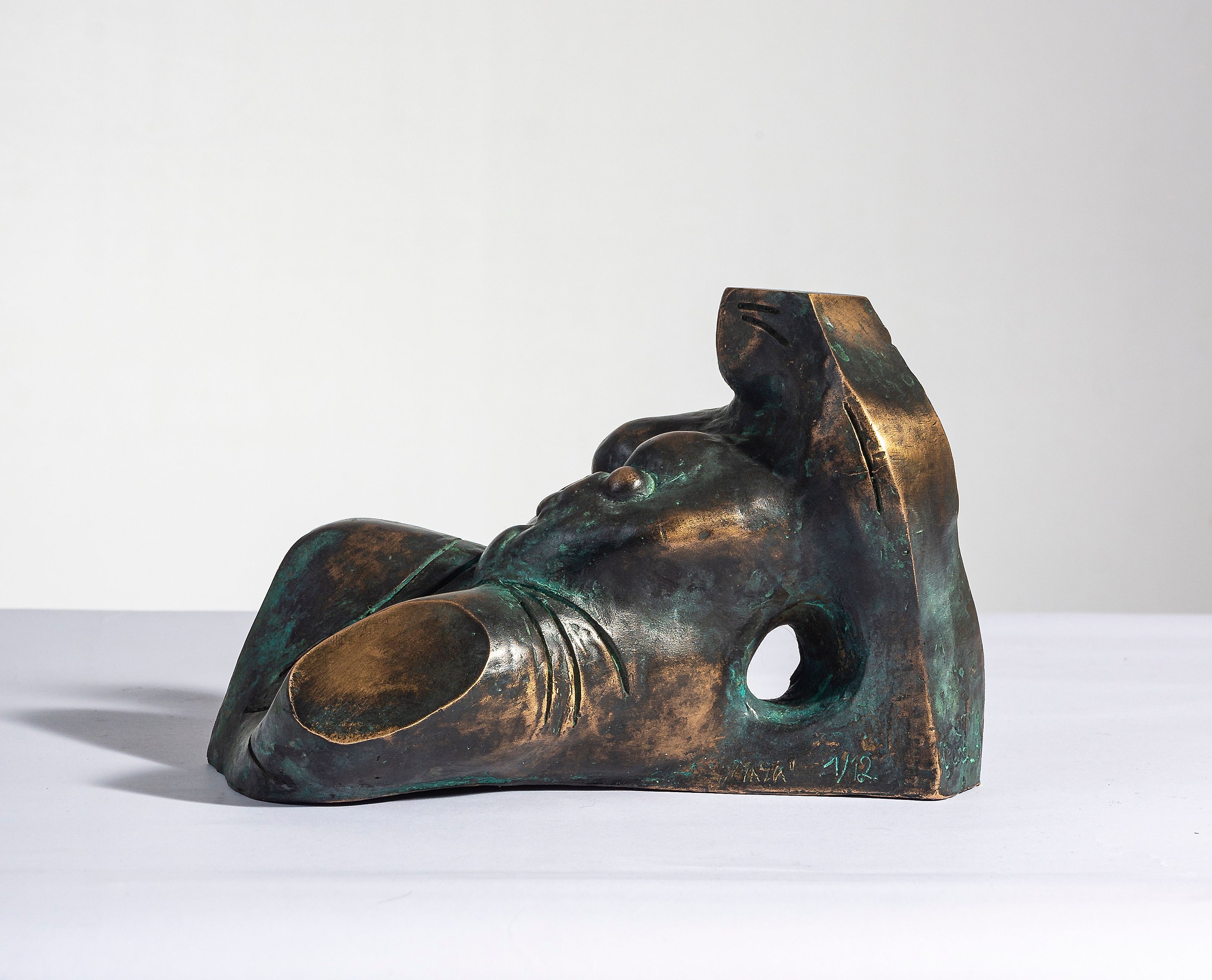 Bronze sculpture “Maria”: a small reclining, simplified figure with a hole through the body, dark bronze with green patina, on a pale surface.
