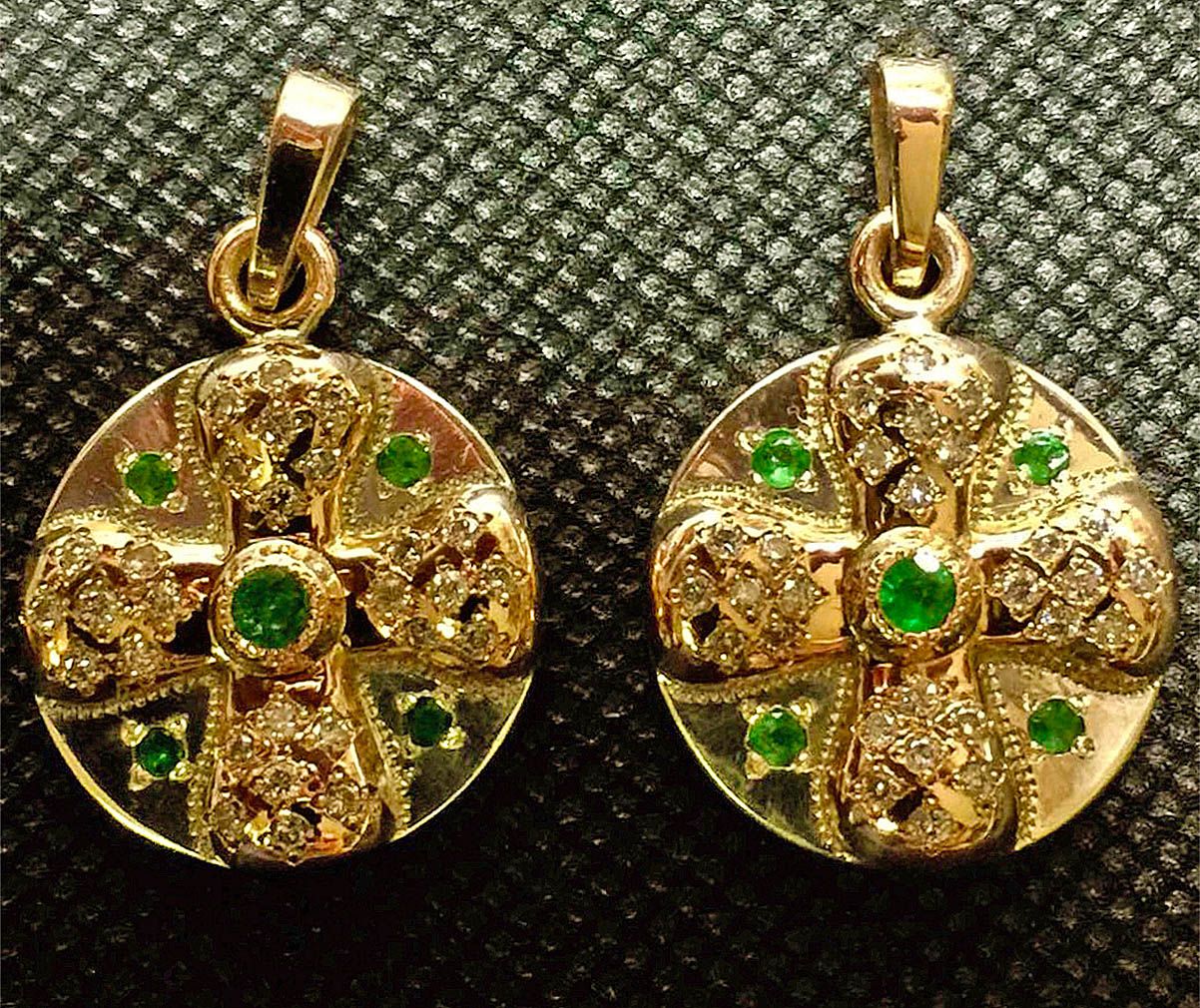 Two gold round pendants, each with a cross-shaped design set with small clear stones and green stones, lying on a dark textured surface.