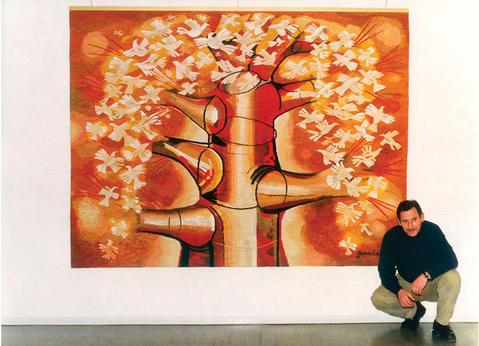 Large tapestry “Radiant Tree” of a pale tree trunk with many white birds in its crown, on a red and orange ground, with the artist crouching beside it in a gallery.