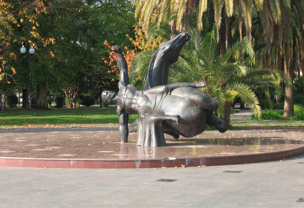 Large bronze sculpture “The Fall and Rise of People Throughout the History”: a falling man and a horse, standing on a round red stone base in a park with palm trees.
