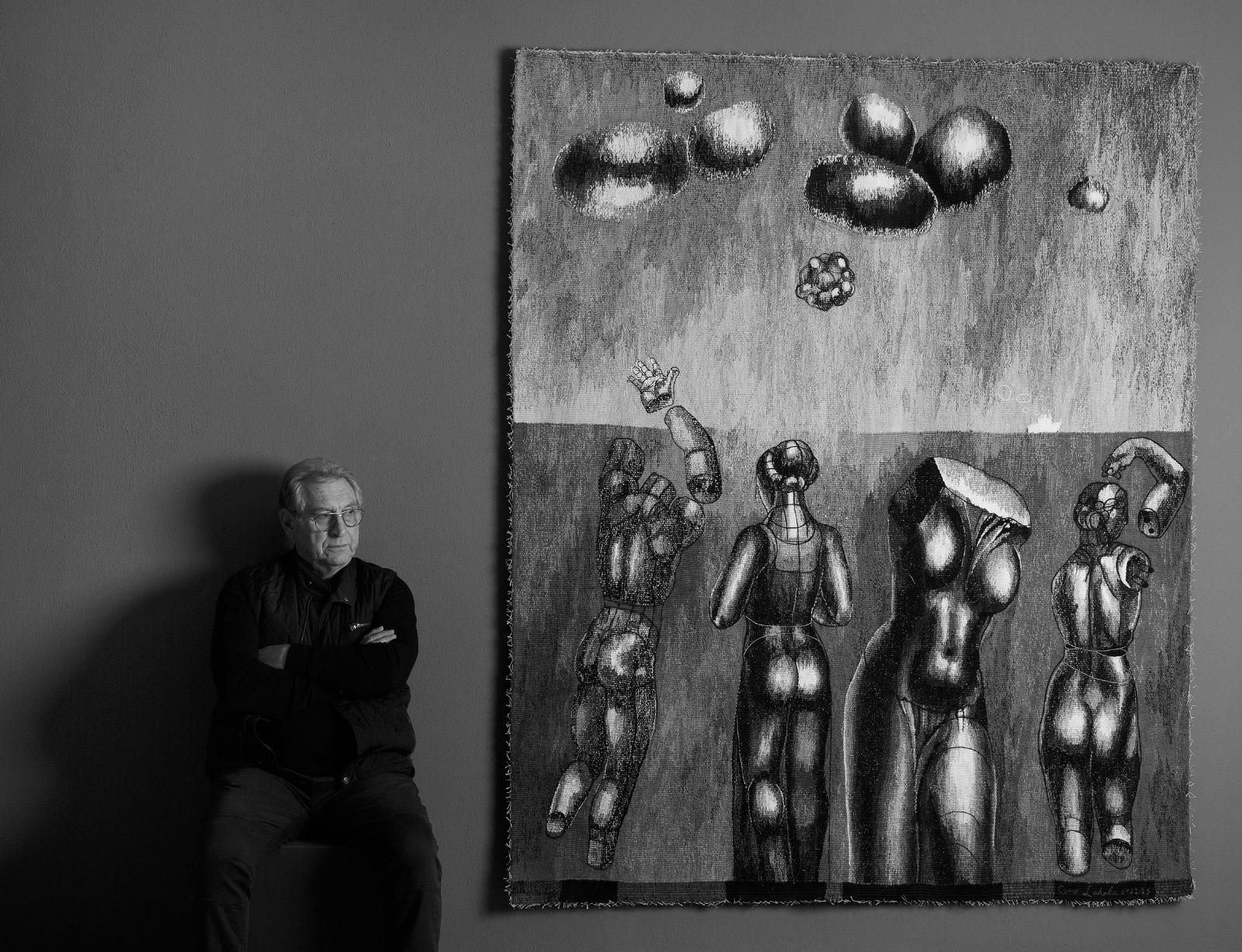 Black-and-white photograph of the tapestry Timelessness hanging on a grey wall: four sculptural female figures and torsos, some with detached arms, stand before a strip of sea with a small white ship, under a sky with dark cloud-like forms. The artist sits beside it with his arms folded.