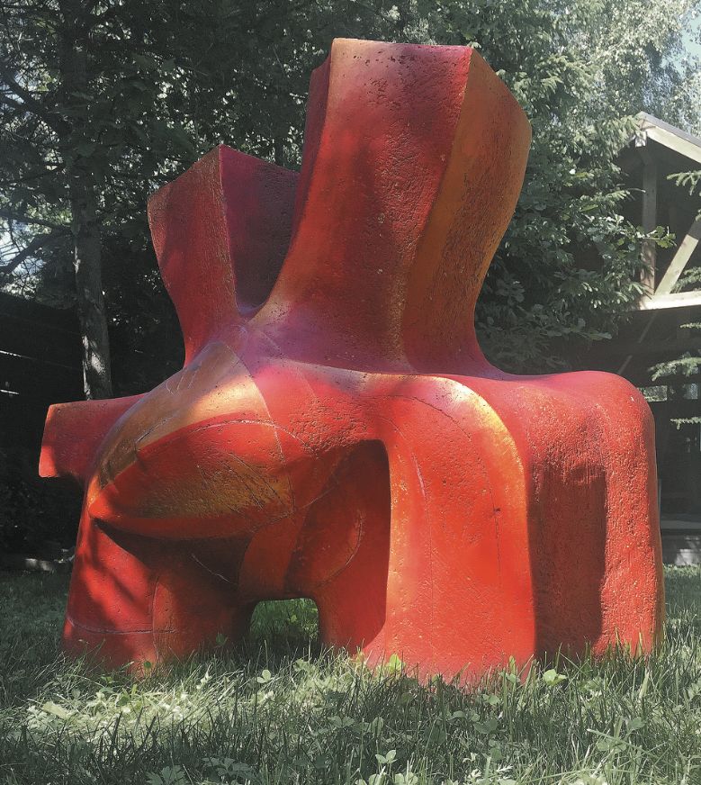 Ceramic sculpture “Yarilo”: a large red, angular abstract form with upright blocky parts, standing on grass among trees.